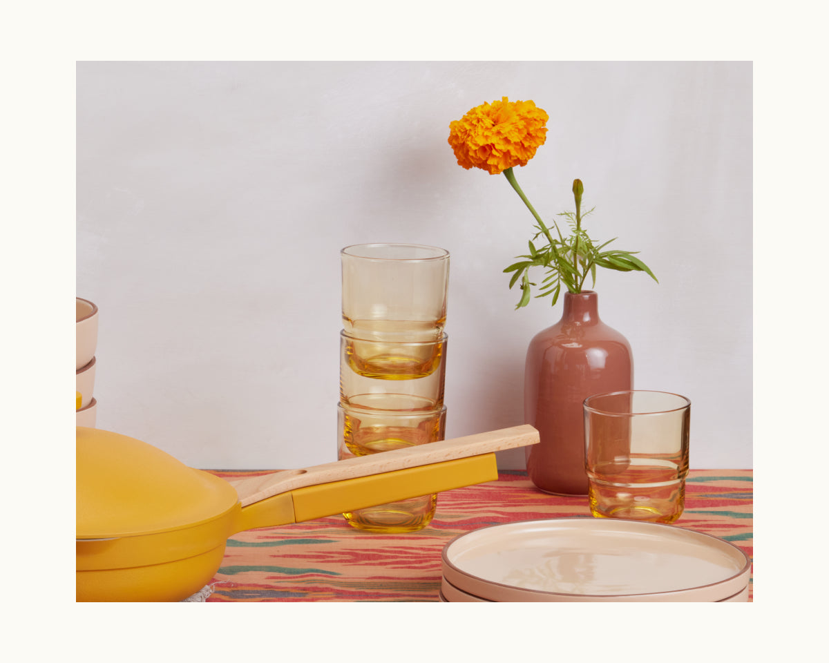 Short Night + Day Glasses, a yellow pan, cream plates, and a single marigold in a mauve vase sit on a patterned tablecloth against a light background.