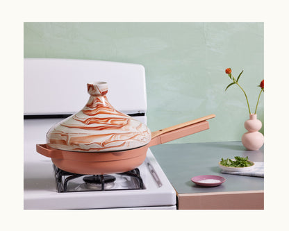 The Tagine, a handcrafted Morocco clay pot, rests on a burner next to a countertop with fresh herbs, salt, and orange flowers—ideal for slow cooking and creating flavorful meals.