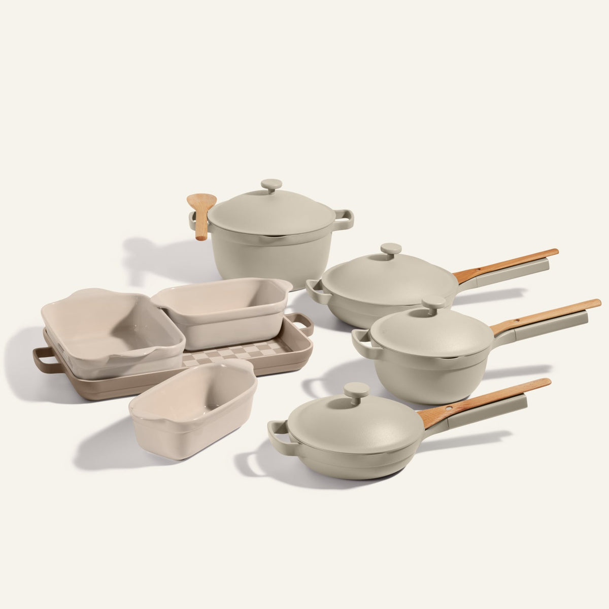 The Cookware + Bakeware Set in light beige features ceramic pots, pans, and baking dishes with wooden handles, including the Perfect Pot and Always Pan, all elegantly arranged on a neutral background.