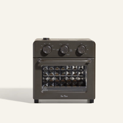 The AU - Wonder Oven is a dark gray countertop toaster oven with three top control dials and a front glass door featuring a diamond-patterned interior. "Our Place" appears at the bottom of the door. Doubles as an air fryer for extra versatility.