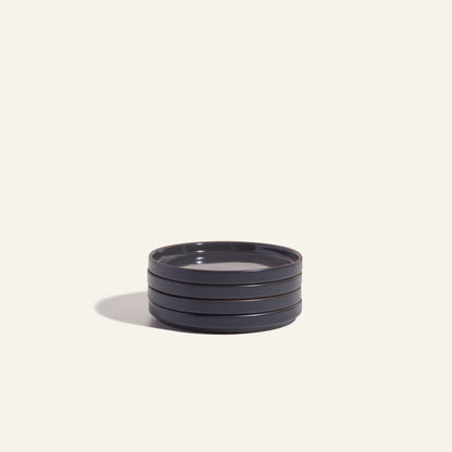 A stack of four round, dark-colored Dessert Plates made from scratch-resistant ceramic sits on a plain light background, casting a shadow to the left.