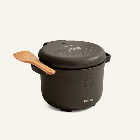 The Our Place Rice Cooker, in black with fuzzy logic and a digital display, sits against a plain background with a wooden spatula on its lid—designed to make perfect rice every time.