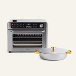 The Wonder Oven Pro, a stainless steel 8-in-1 countertop oven with a digital display, sits next to the Titanium Braiser Pro—a silver and gold lidded braiser—on a light background.