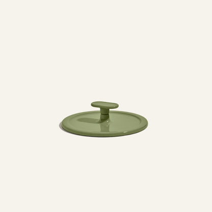 A green circular cast iron Grill Press with a central handle, displayed on a plain light background, designed for even heat distribution.